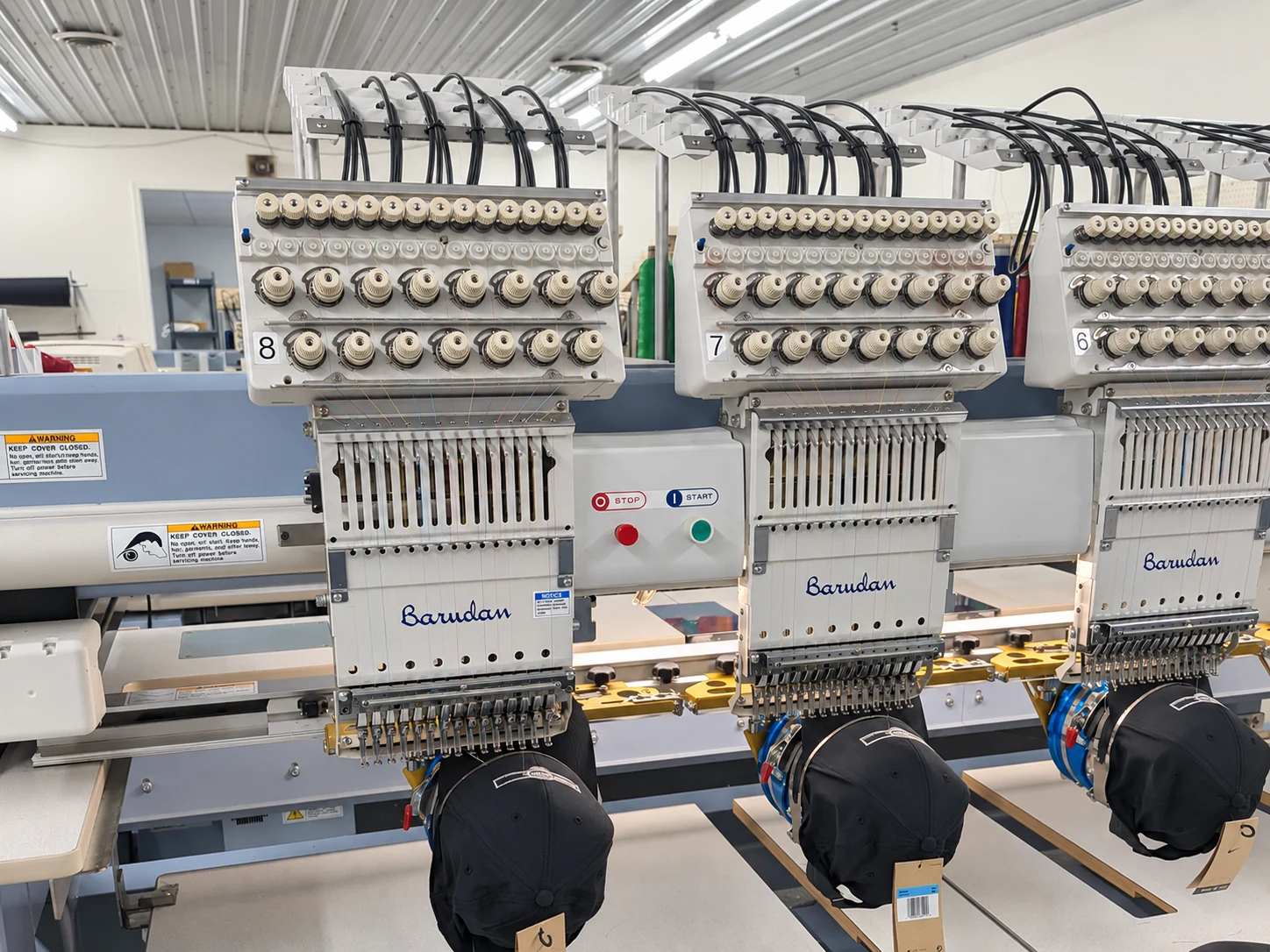 Edited photo: Black caps mounted along a line of embroidery machine heads at Del Print