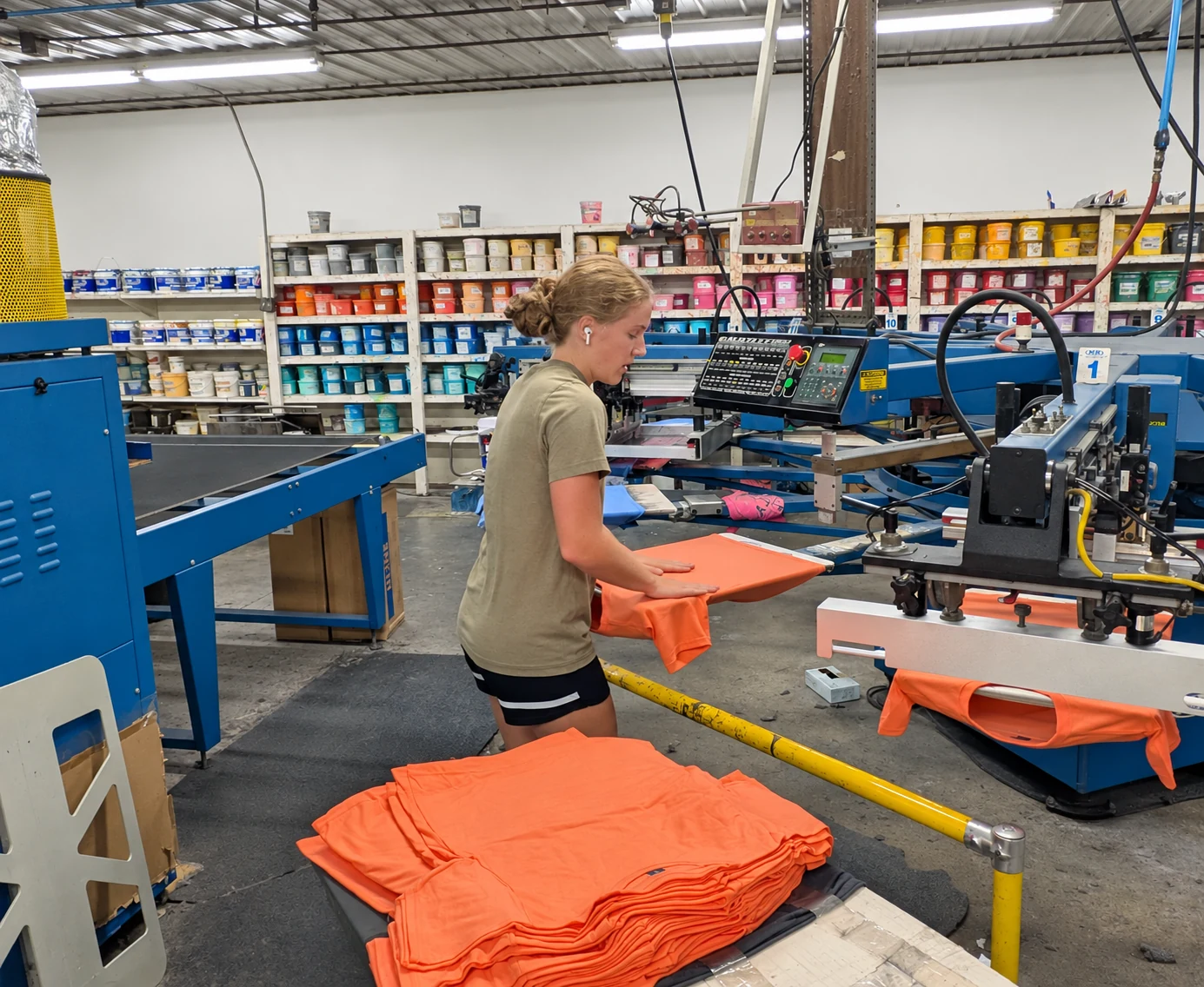 Edited photo: Del Print employee handling a stack of orange shirts beside the press